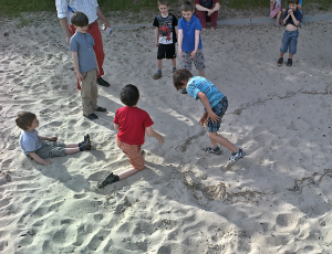 einige Kinder befinden sich mit Spielleiter Robert Mingau in einem Sandkasten, dort wurde mit einem Kreis aus Wasser eine Kampfarena markiert