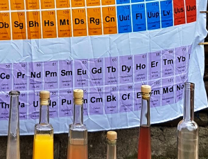 Dekoration für Chemieexperimente - Flaschen mit bunten Flüssigkeiten vor einem Periodensystem-Vorhang