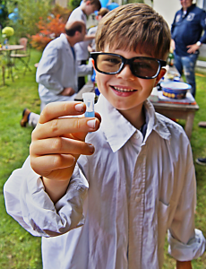 Ein Junge mit Laborkittel und Schutzbrille präsentiert begeistert ein Mini-Reagenzgefäß
