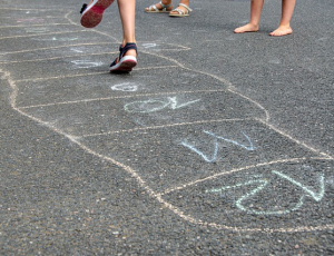 Auf dem Asphalt sieht man Kreidefelder mit Zahlen für ein Hüpfspiel, im Hintergrund sind die Füße hüpfender Kinder zu erkennen