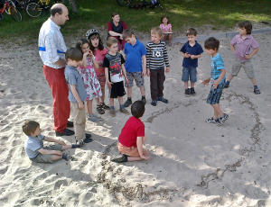 Kinderolympiade: in einem Sandkasten wurde ein Ring für einen Ringkampf markiert, zwei Kinder sind darin und bereiten sich auf den Kampf vor, eine Kindergruppe mit Spielleiter Robert steht rund um
den Ring
