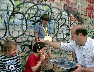 Kinderolympiade - Siegerehrung: Der Spielleiter hängt einem der drei Sieger eine Medaille um den Hals. Im Hintergrund ist eine mit vielen Graffitis besprayte Mauer zu sehen.