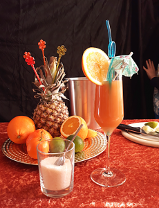 Stilleben in Orange: Dekorative Früchte, Zuckerglas und gefülltes Cocktailglas auf orangebrauner Tischdecke