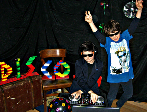 Kinderdisco mit einem DJ und einem tanzenden Jungen. Die Buchstaben DISCO, aus bunten Legosteinen gebaut und angeleuchtet, bilden die Dekoration.