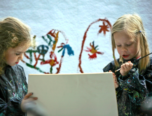 zwei etwa fünfjährige Künstlerinnen sind in die Arbeit vertieft, ein Ausschnitt ihres expressiven Gemäldes ist erkennbar
