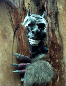 ein Teufel mit sehr gruseliger Maske schaut aus einem Baumversteck heraus