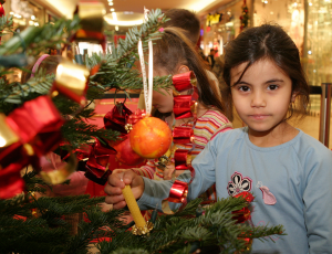 Ein Mädchen steht neben einem reich geschmückten Weihnachtsbaum und blickt nachdenklich in die Kamera