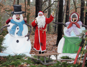 Der Weihnachtsmann steht winkend zwischen zwei lebensgroßen Schneemann-Figuren, die als Fotokulisse im Wald aufgebaut wurden