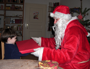 Der Weihnachtsmann übergibt einem etwa dreijährigen Kind in einer Wohnung ein rotes Geschenkpäckchen